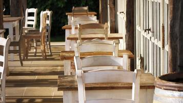 A row of white wooden chairs and tables under a covered area with a wooden barrel.