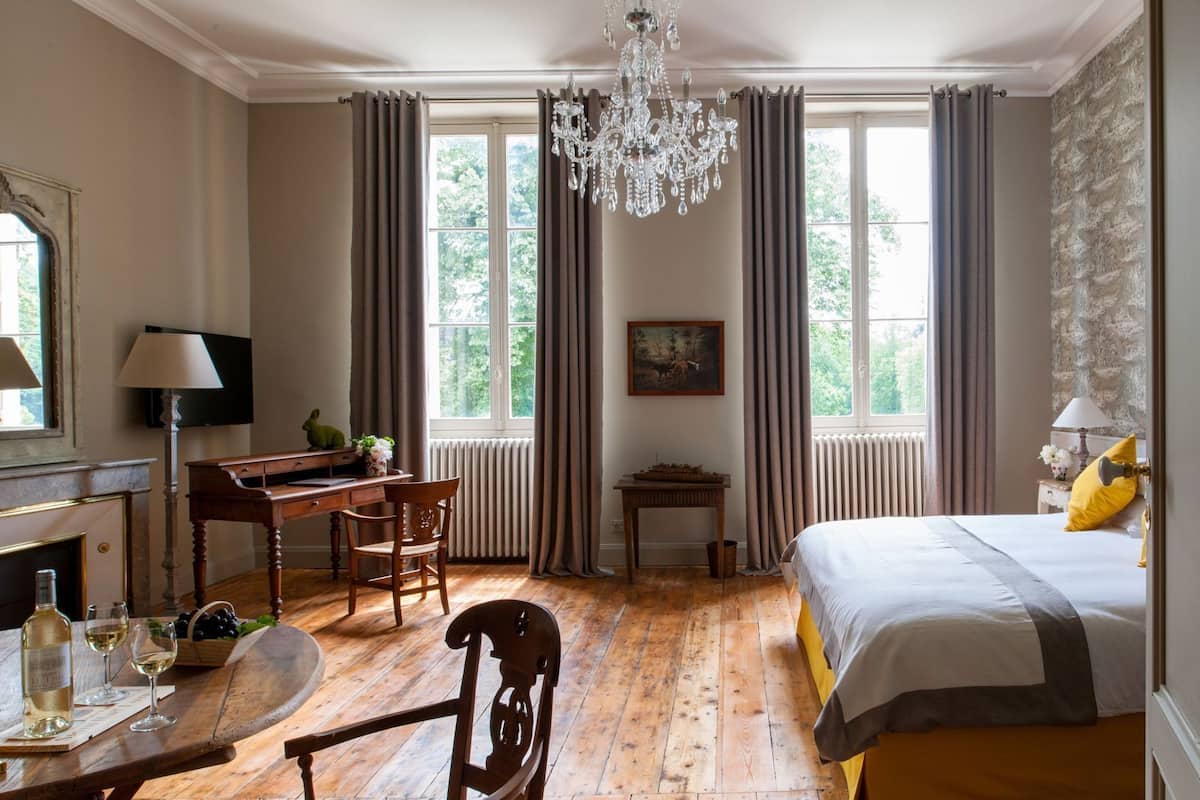 Une pièce comprenant un lit, un bureau, une chaise, un lustre et de grandes fenêtres avec des rideaux.