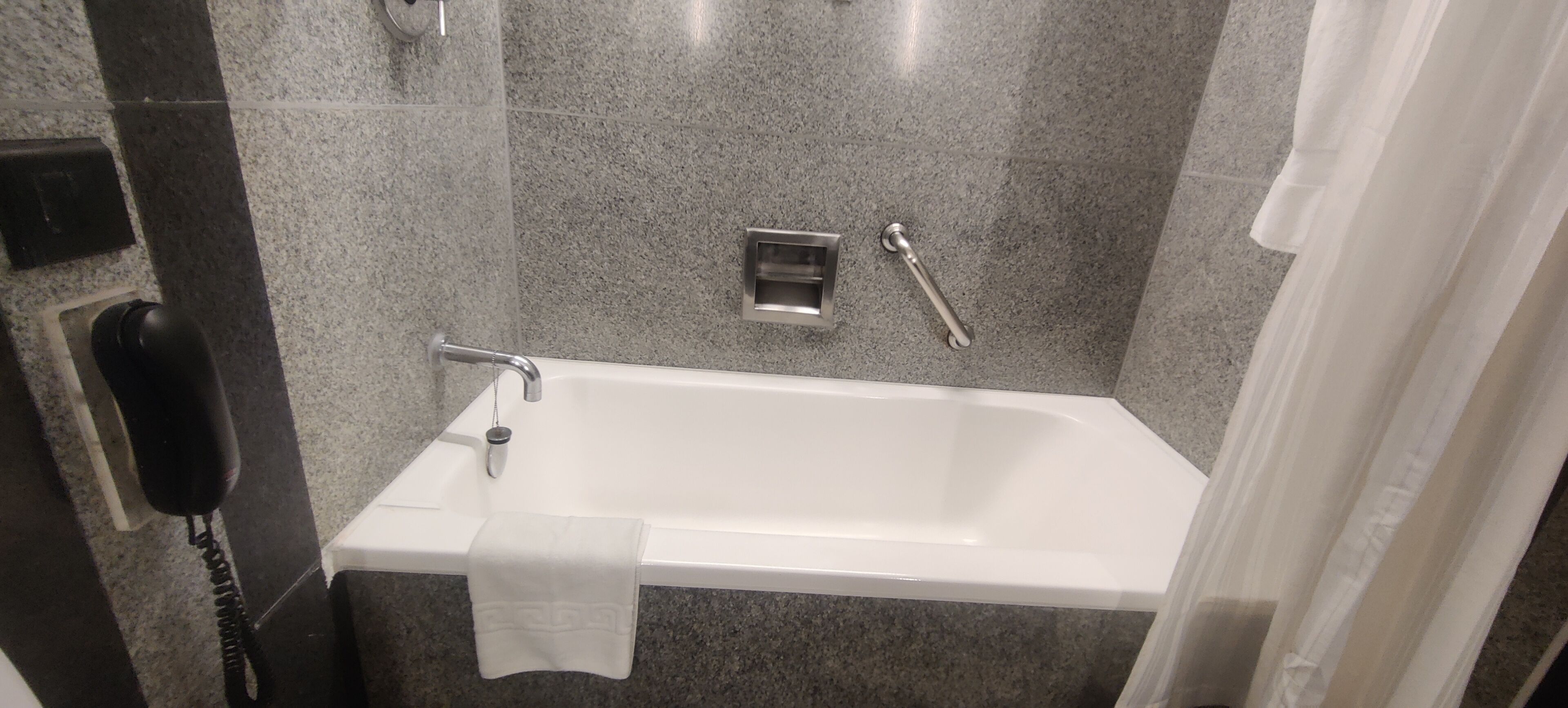 A modern bathroom with a white bathtub, a handheld showerhead, a towel, and a hand dryer.