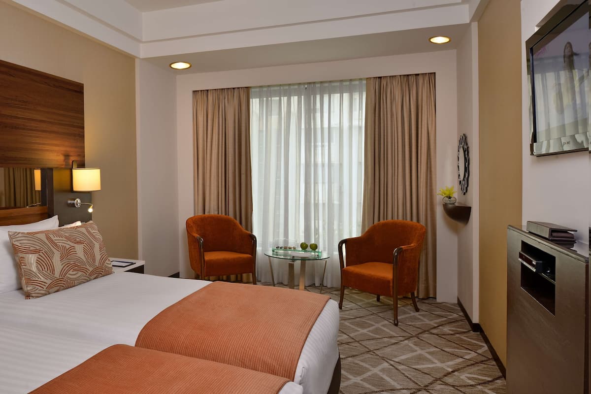 Quarto de hotel com uma cama grande, duas poltronas, uma mesinha de centro com um vaso e uma televisão.