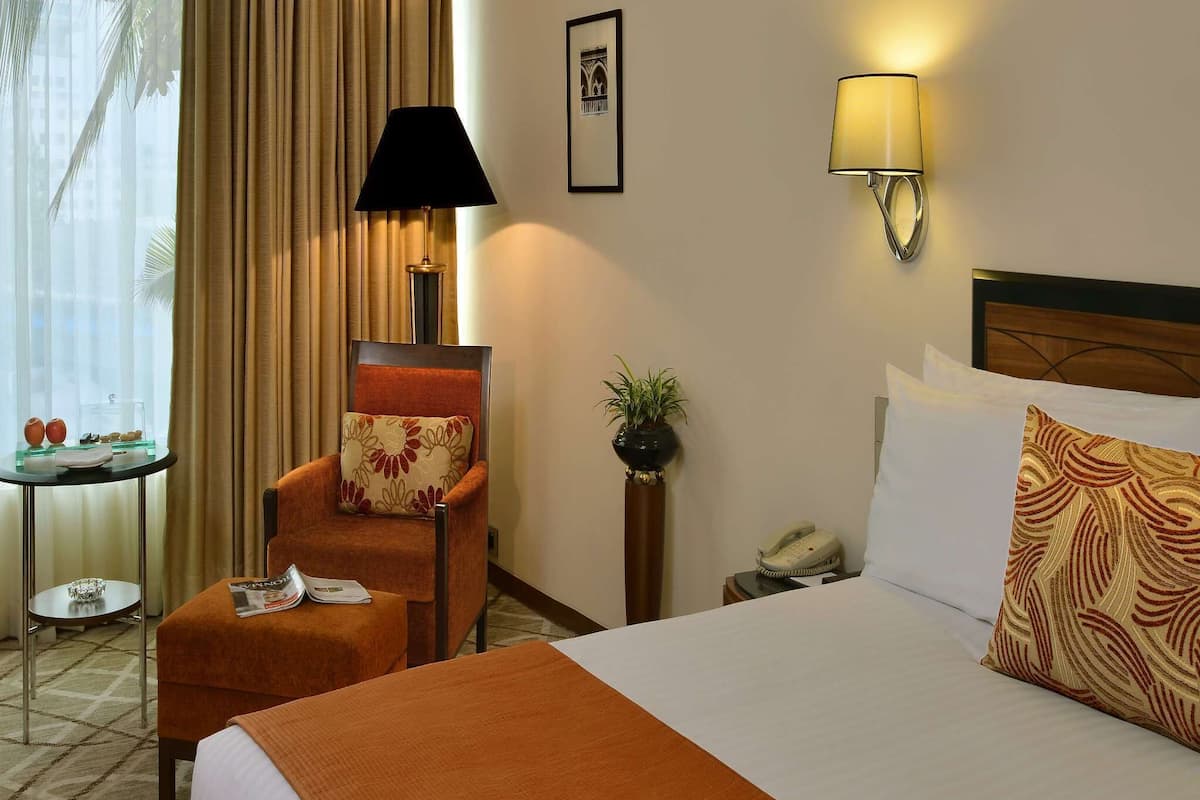 Quarto de hotel com uma cama, uma cadeira, uma mesinha com abajur, uma planta em vaso e um quadro na parede.