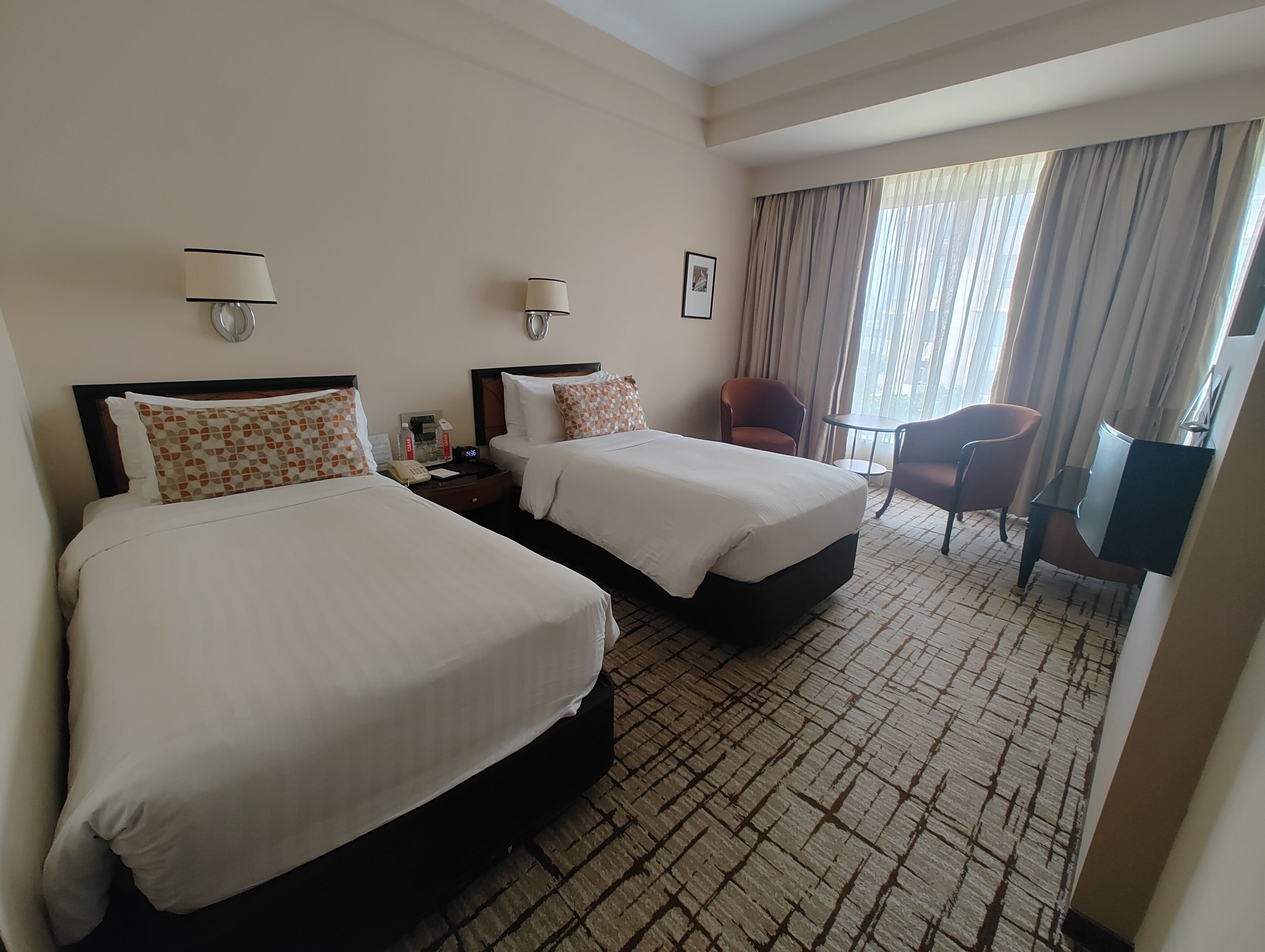 A hotel room with two beds, a desk, a chair, and a large window with curtains.