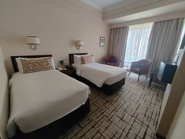A hotel room with two beds, a desk, a chair, and a large window with curtains.