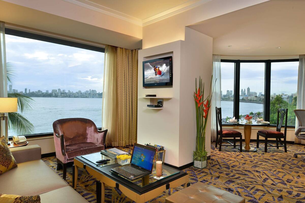Quarto de hotel com uma janela ampla que oferece vista da cidade, uma mesa com um laptop, um sofá e um espaço para refeições.