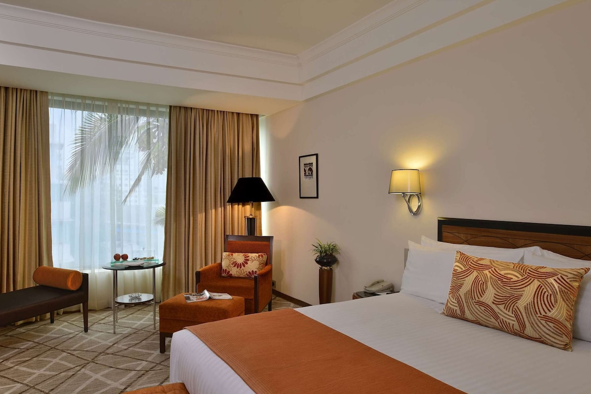 A hotel room with a bed, a desk, a chair, a lamp, and a view of palm trees.