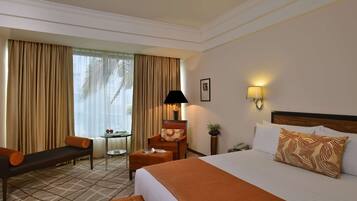A hotel room with a bed, a desk, a chair, a lamp, and a view of palm trees.