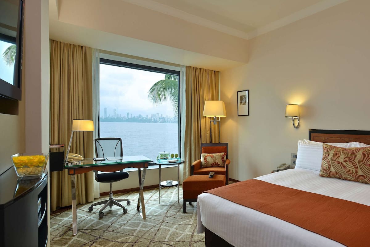 Quarto de hotel com uma cama grande, uma secretária com cadeira, janela com cortinas e vista para o horizonte urbano.