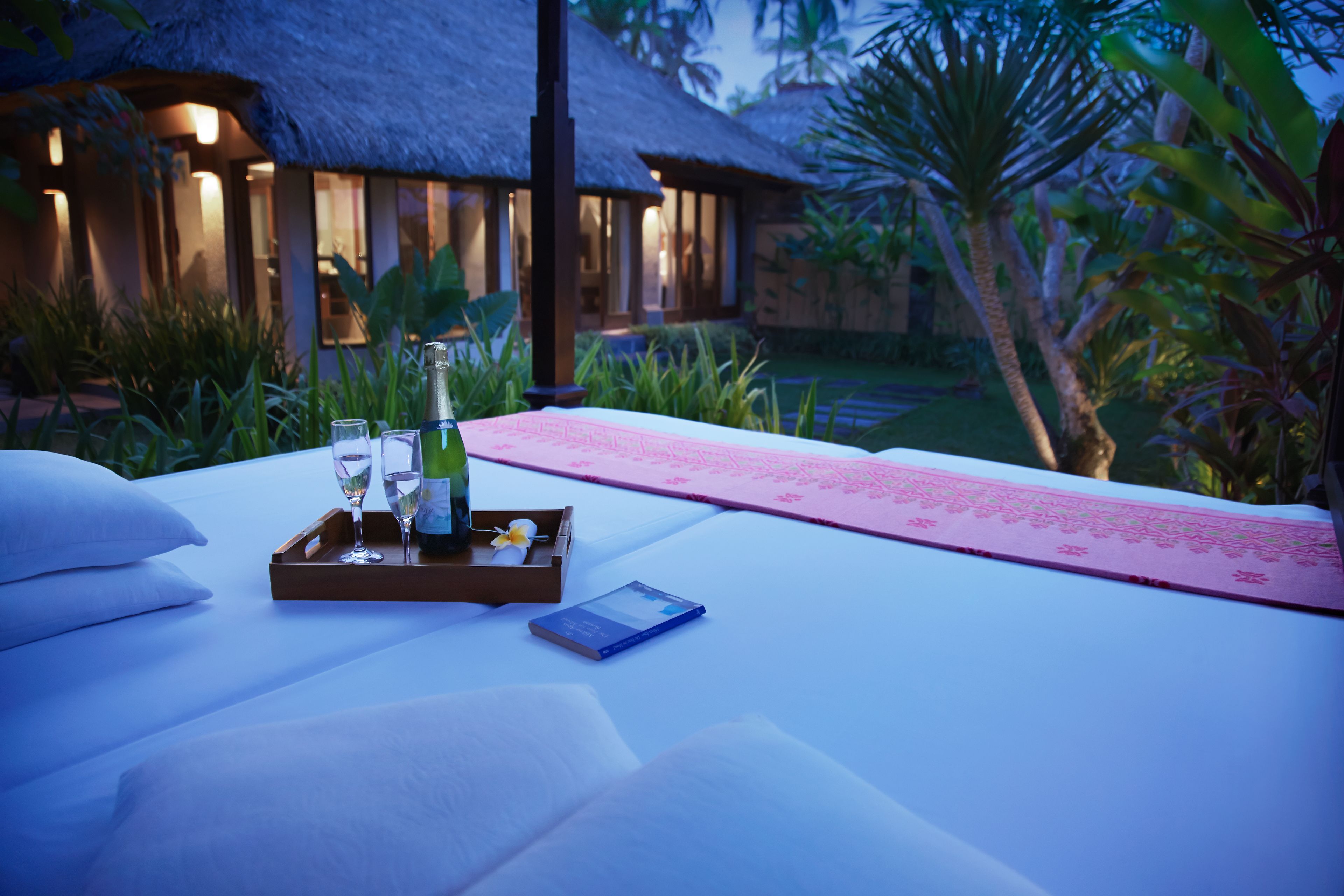 A bed with a tray containing a bottle, glasses, and a book, set against a backdrop of a thatched-roof building and lush greenery.