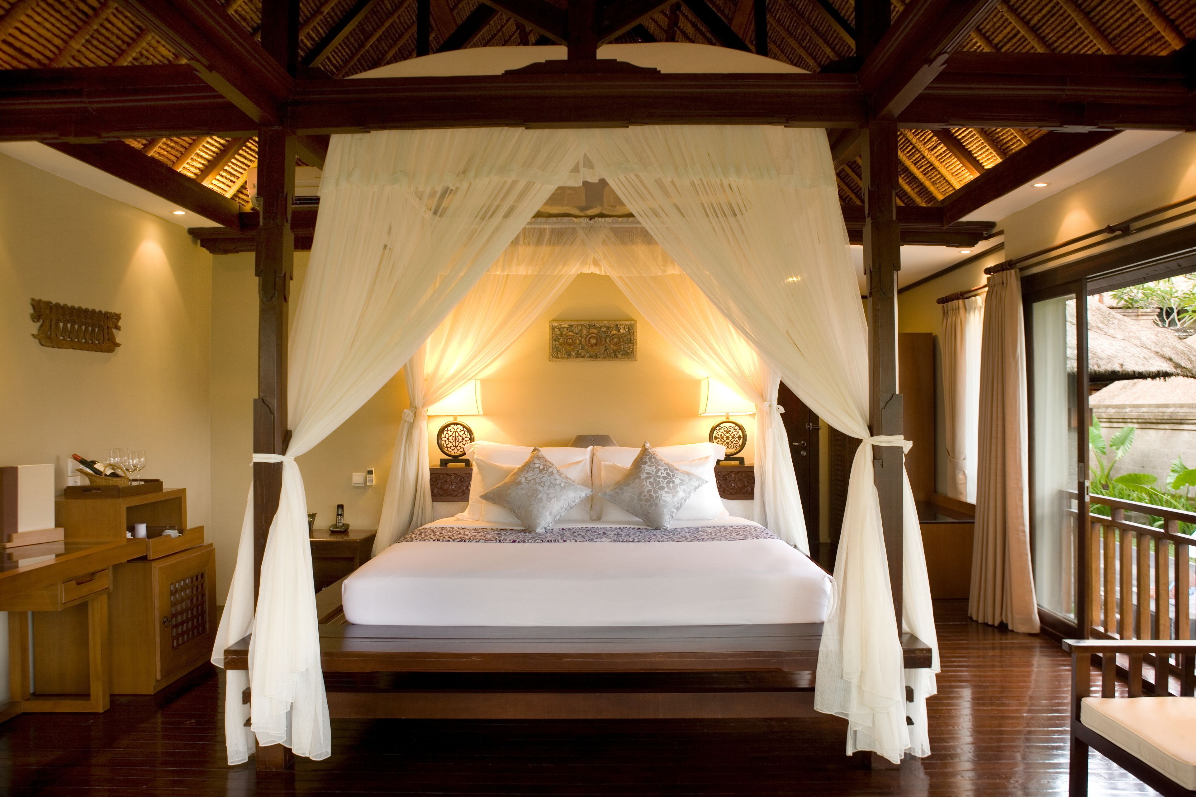 A four-poster canopy bed with white linens and decorative pillows, wooden furniture, and a view of greenery outside.