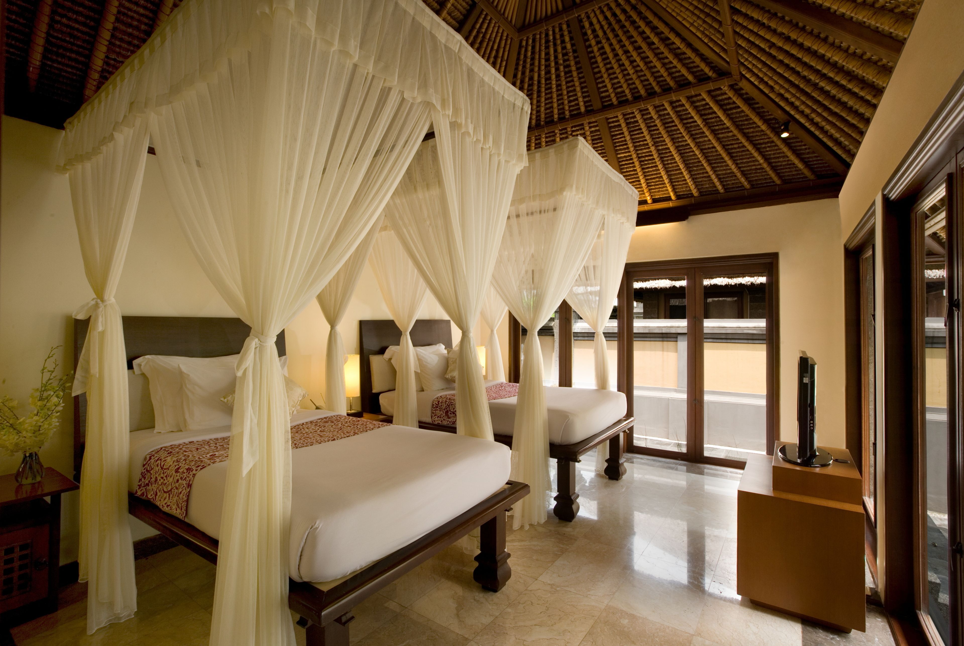 A hotel room with two beds, each with a canopy, a wooden headboard, and a bedside table with a lamp.