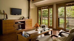 A living room with a wooden cabinet, a television, a laptop, and a window with a view of greenery.
