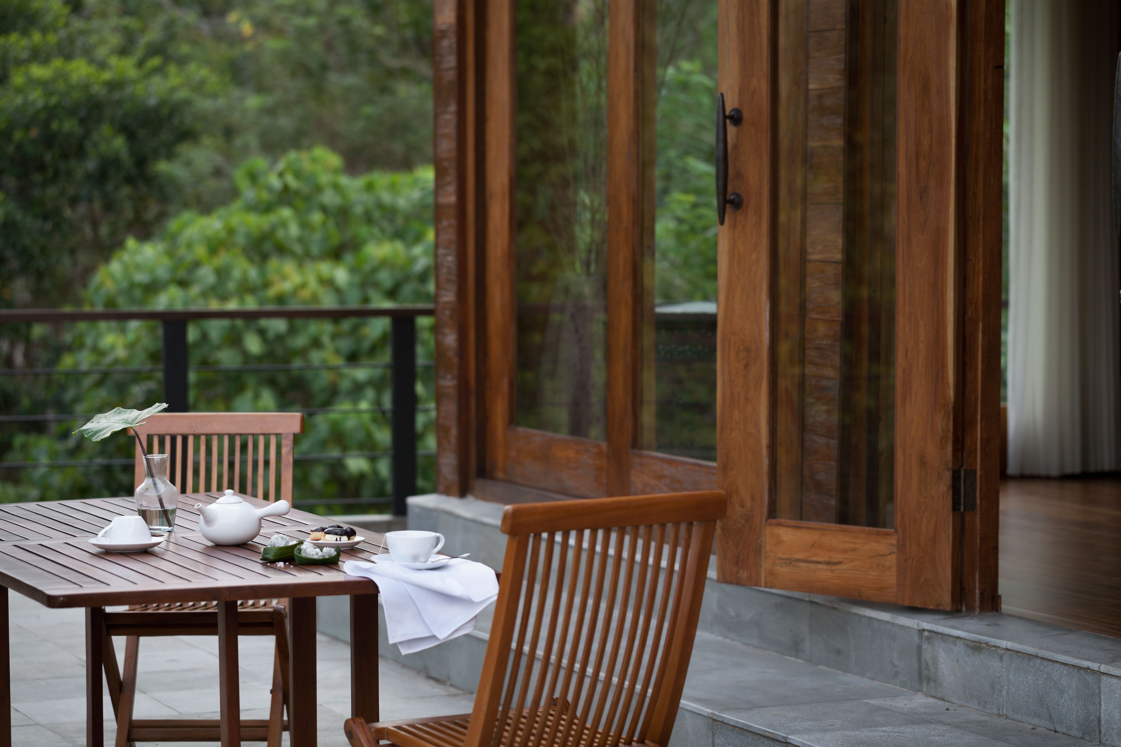 A wooden table with a teapot, cups, and a small vase on a balcony with a view of greenery.