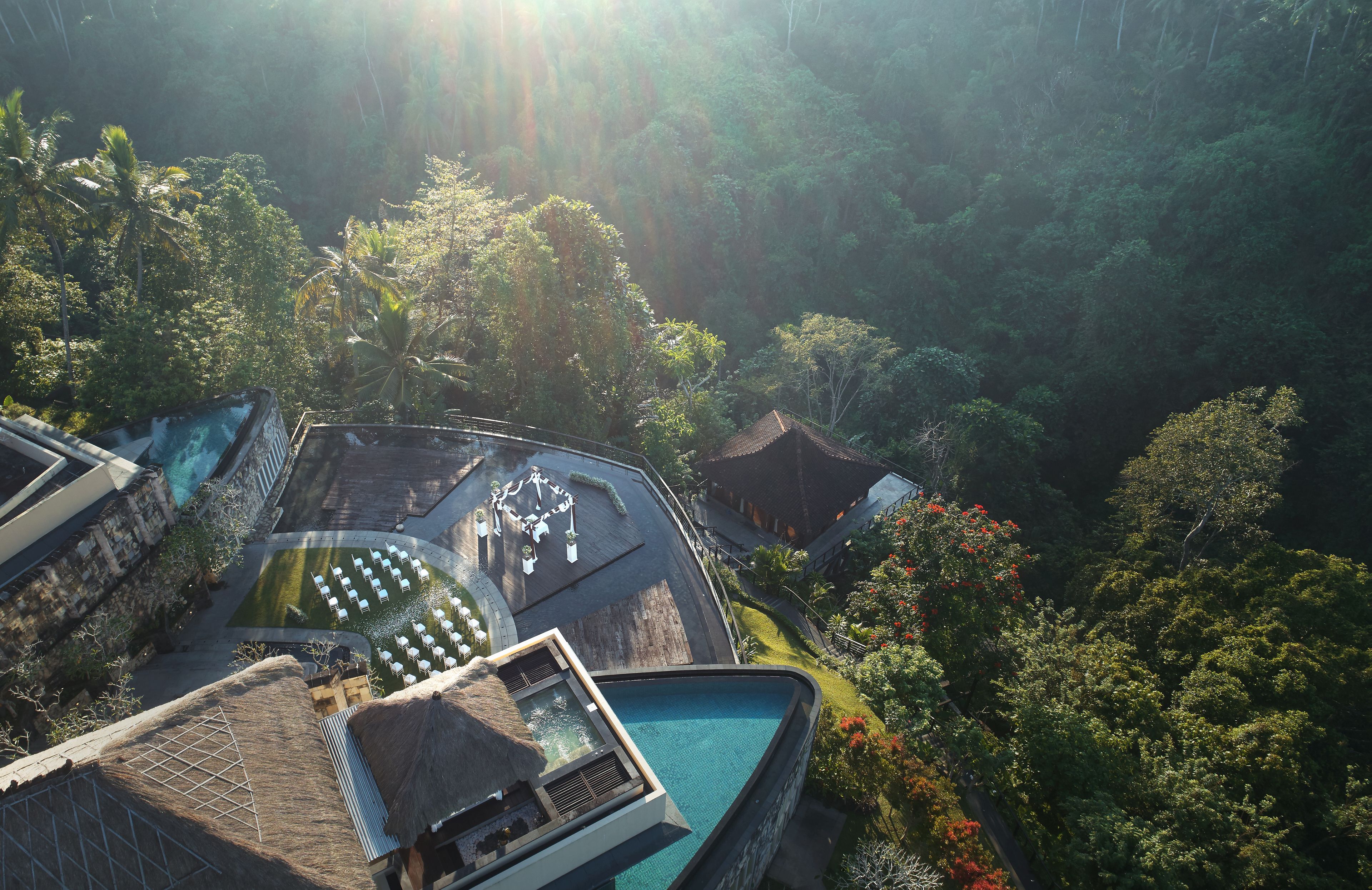 An aerial view of a resort with a swimming pool, lounge chairs, and a thatched-roof structure surrounded by lush greenery.