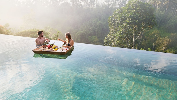 A couple dining on a floating platform in a clear pool with a tropical forest backdrop.