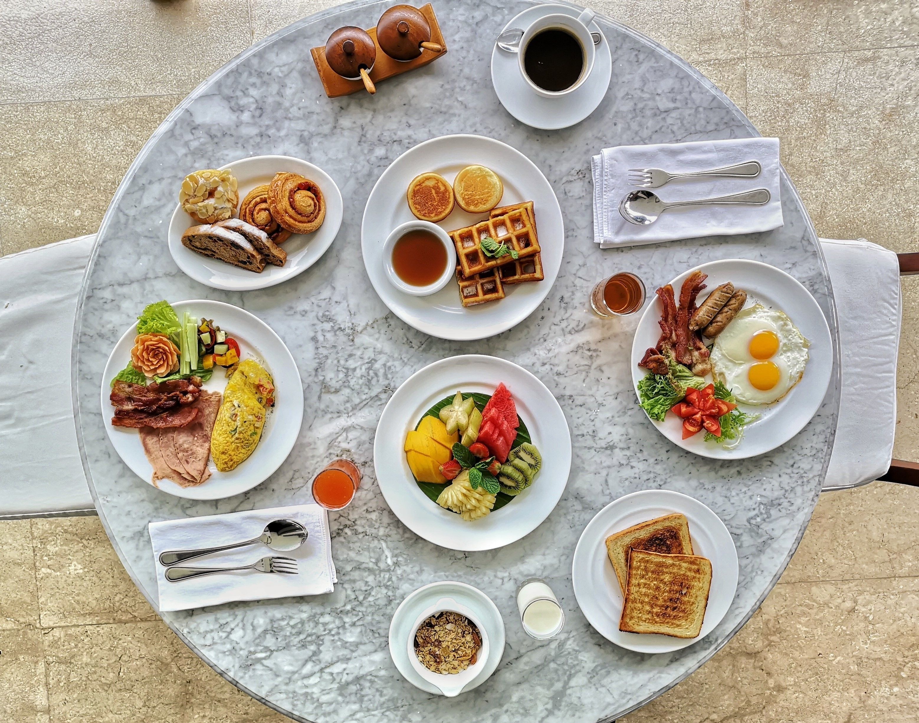 A breakfast spread on a marble table with various dishes including waffles, eggs, toast, and fruit.