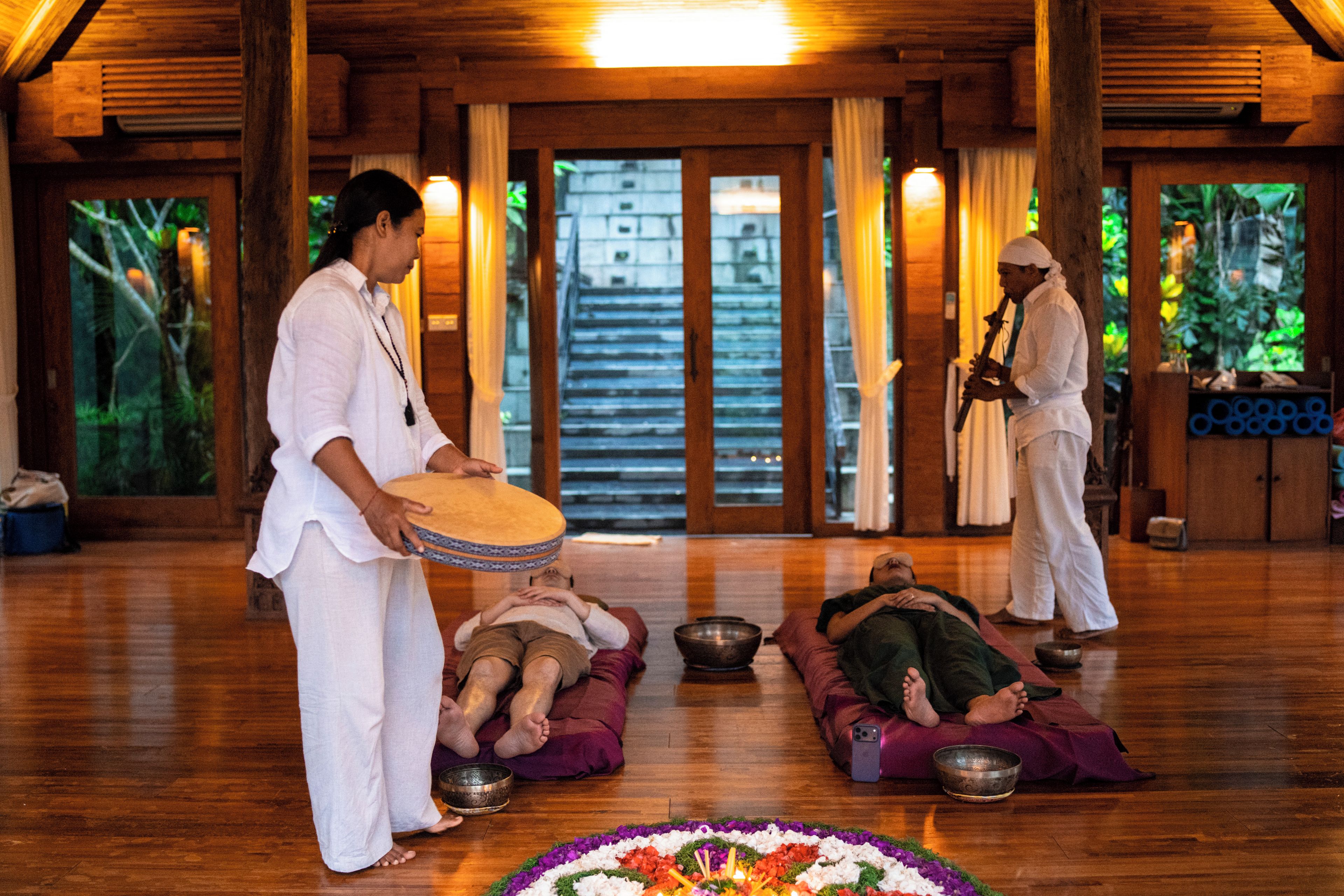 A traditional ceremony with participants seated on cushions, a person holding a drum, and a person playing a flute.
