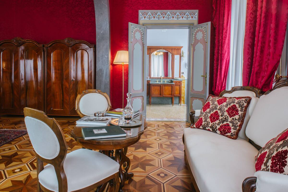 Una habitación con cabecera de madera, sofá blanco con cojines estampados, una mesa de madera con lámpara y piso con diseño.