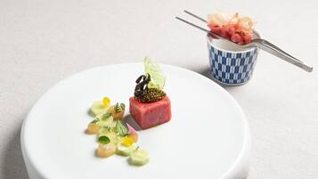 Ein weißer Teller mit verschiedenen Speisen, darunter ein Stück rohes Fleisch, Gemüse und eine kleine, blau-weiße Tasse mit einem Löffel.
