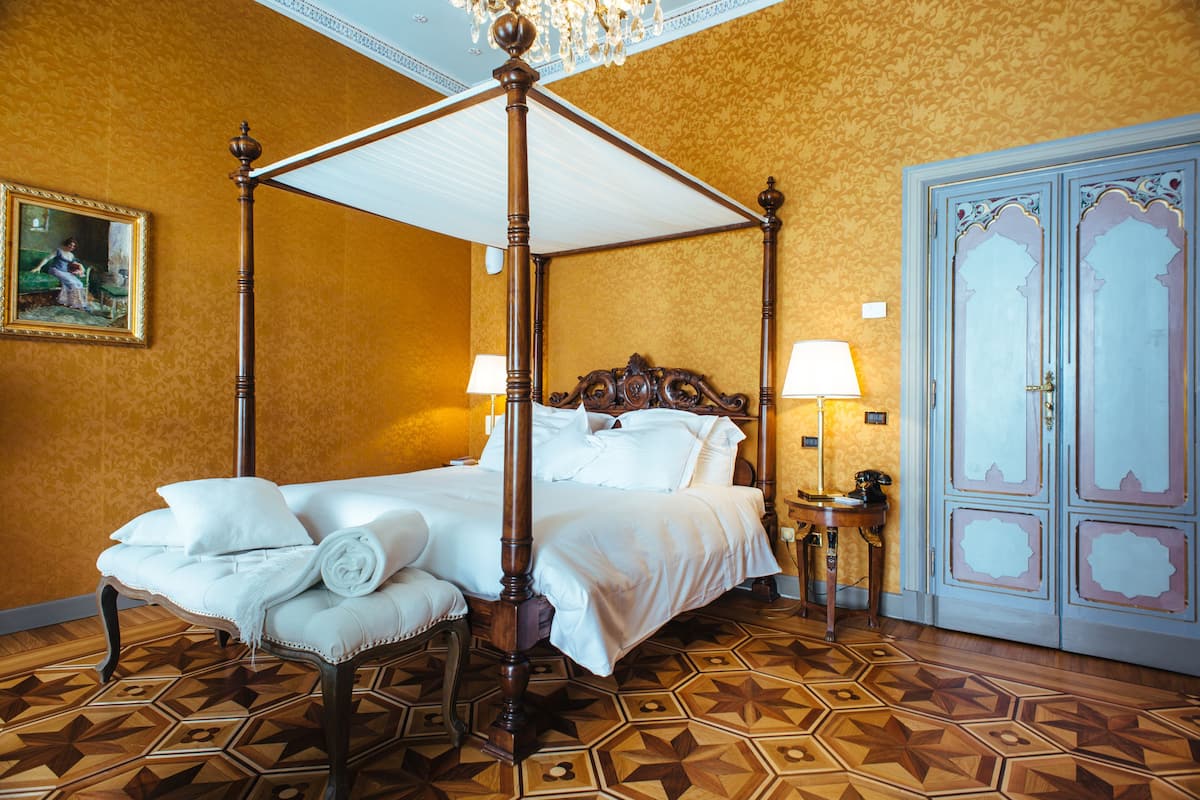 Una cama con dosel con ropa de cama blanca, una toalla enrollada, una mesita de noche con una lámpara y una puerta grande y decorada.