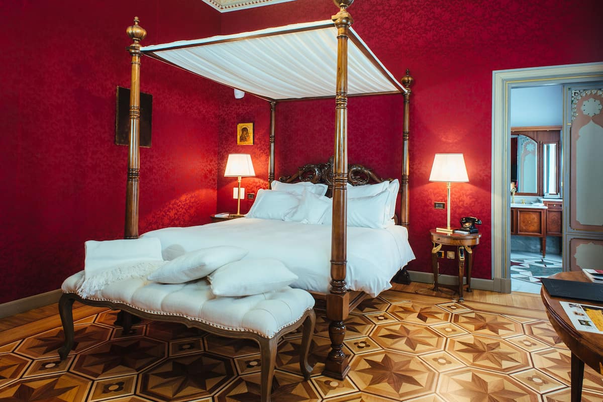 Una cama con dosel cubierta con sábanas blancas, una pared roja y un suelo con estampado geométrico.