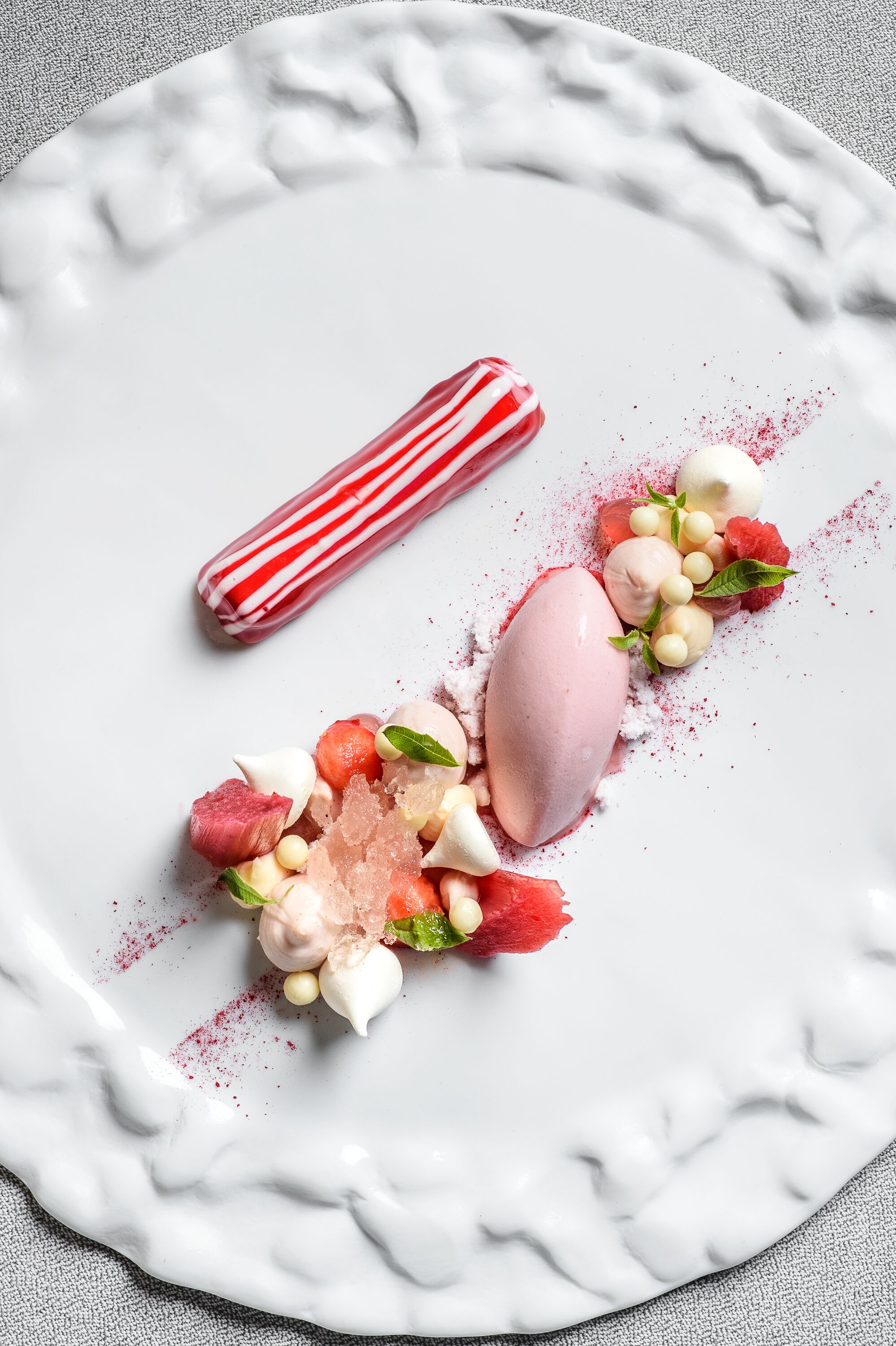 A dessert plate with a white marble pattern, featuring a pink ice cream scoop, a striped candy, and various garnishes including berries and mint leaves.