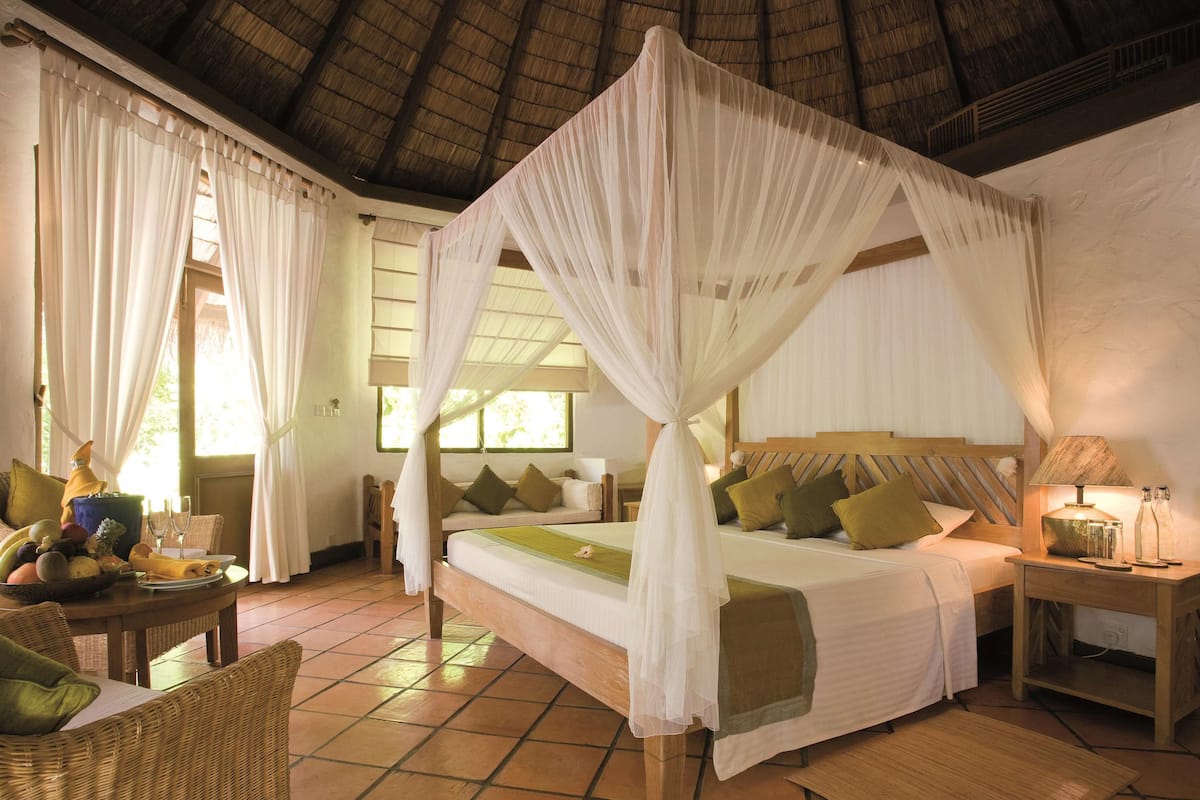 A four-poster bed with a canopy, a dining table with fruit and glasses, wicker chairs, and a wooden nightstand with a lamp.