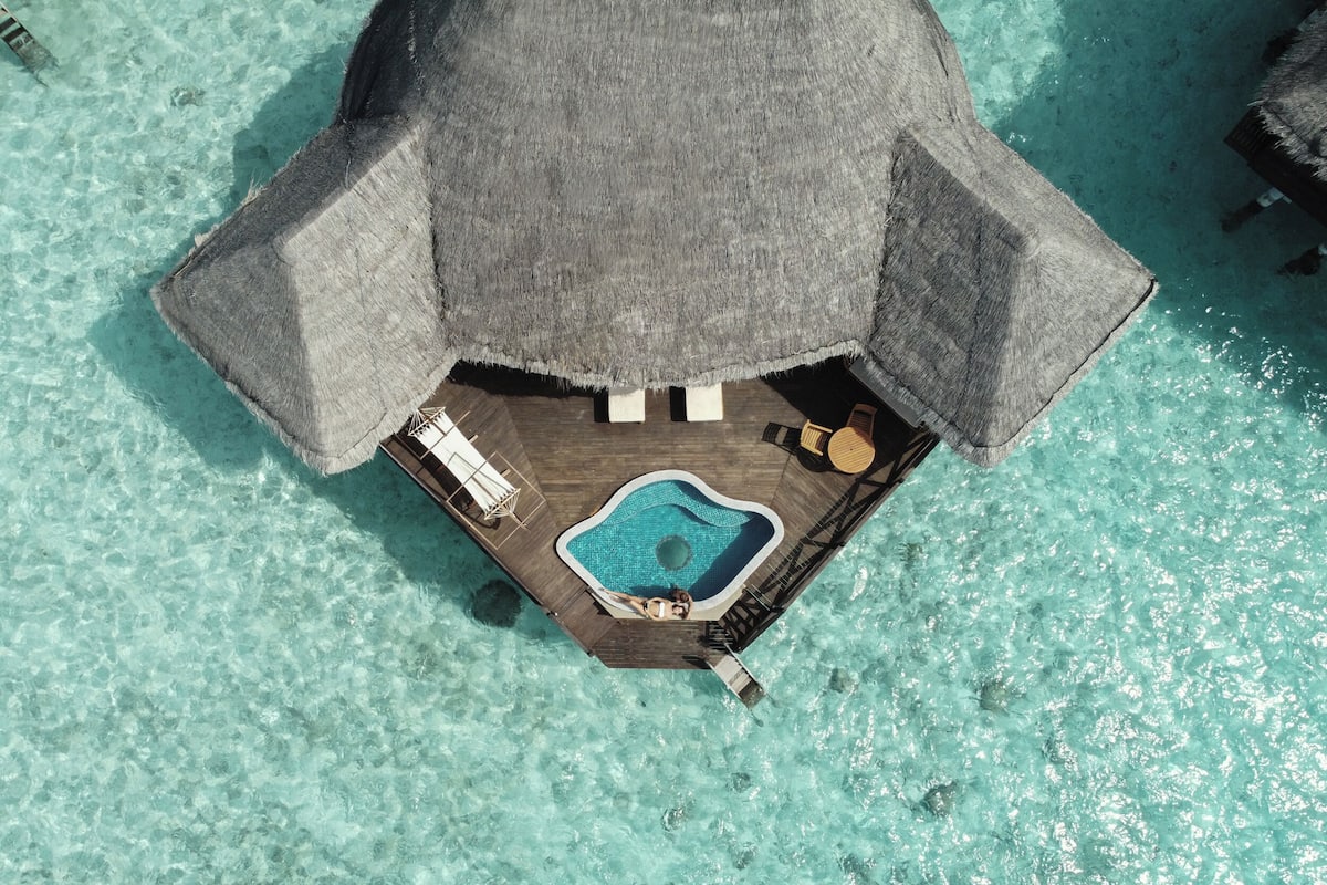 An overwater bungalow with a thatched roof, a small pool, and a wooden deck with lounge chairs.
