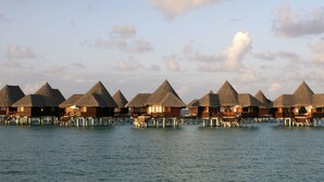 Des bungalows sur pilotis, avec toits de chaume, s’étendent au-dessus d’une mer calme.