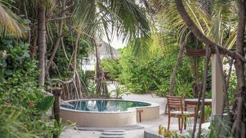 A circular swimming pool surrounded by lush greenery and a wooden deck with chairs.