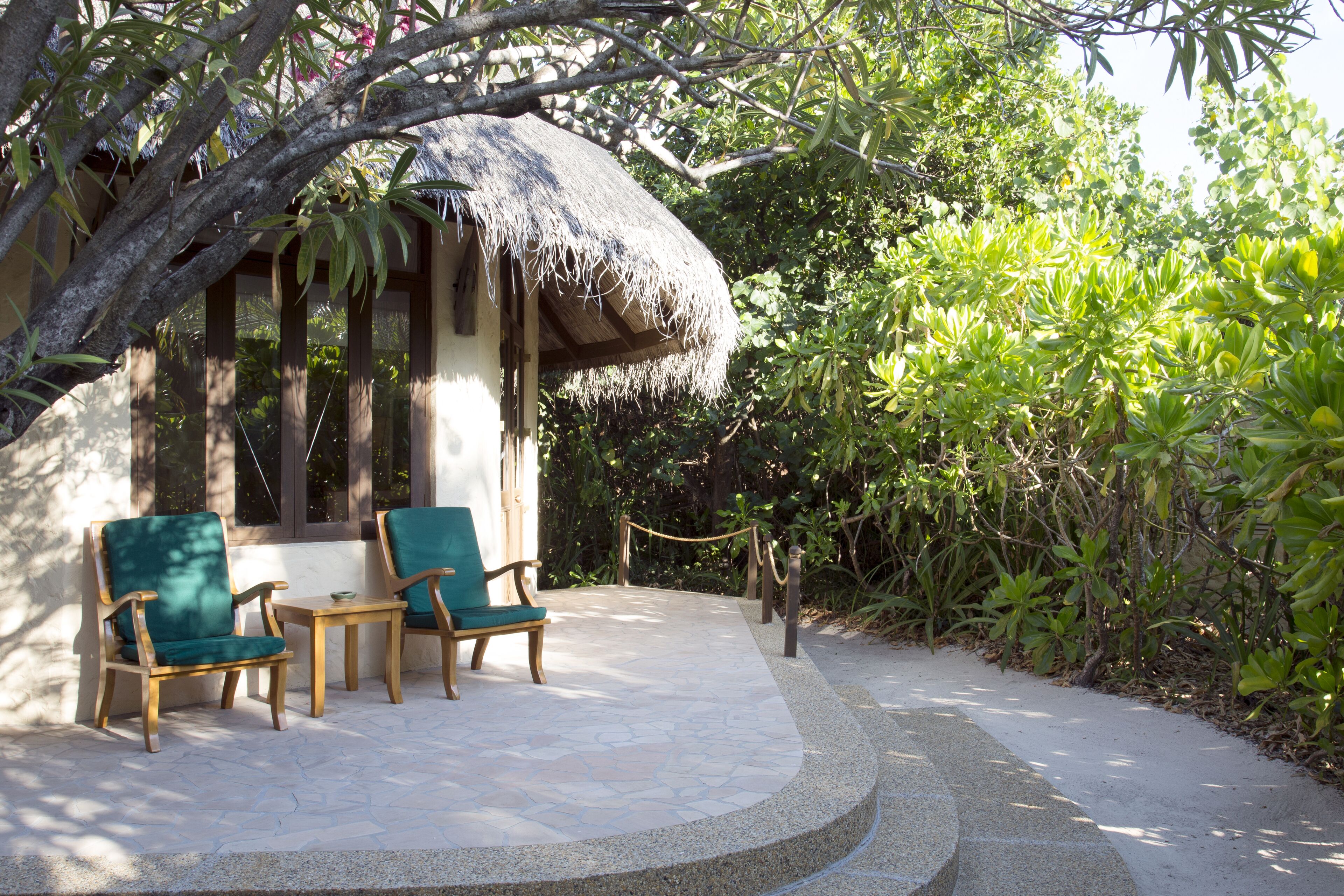 A thatched-roof structure with two wooden chairs and a small table, surrounded by lush greenery.
