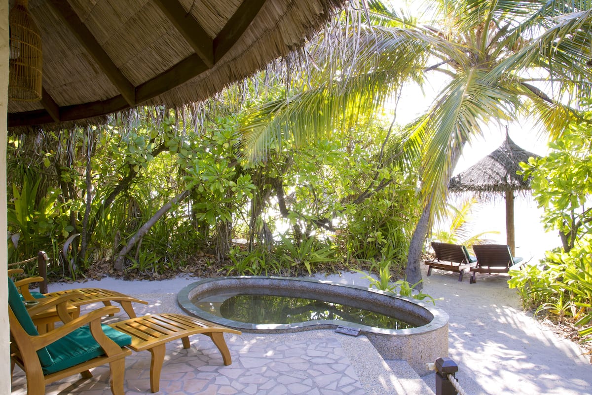 A tropical outdoor area with a thatched-roof structure, a small pool, wooden lounge chairs, and lush greenery.