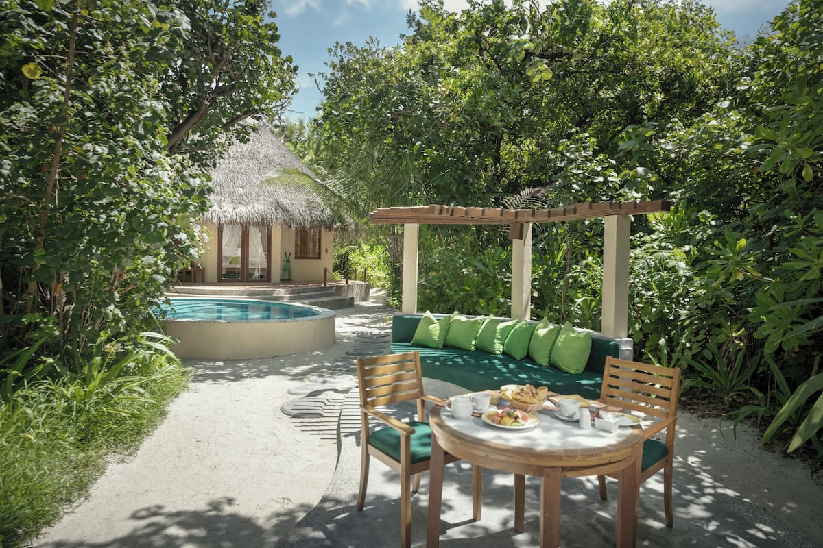 A pool area with a thatched-roof hut, a wooden pergola, and a green sofa with cushions.