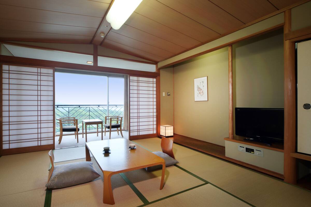 A traditional Japanese room with tatami flooring, a low table with cushions, a television on a stand, and a sliding door leading to a balcony.
