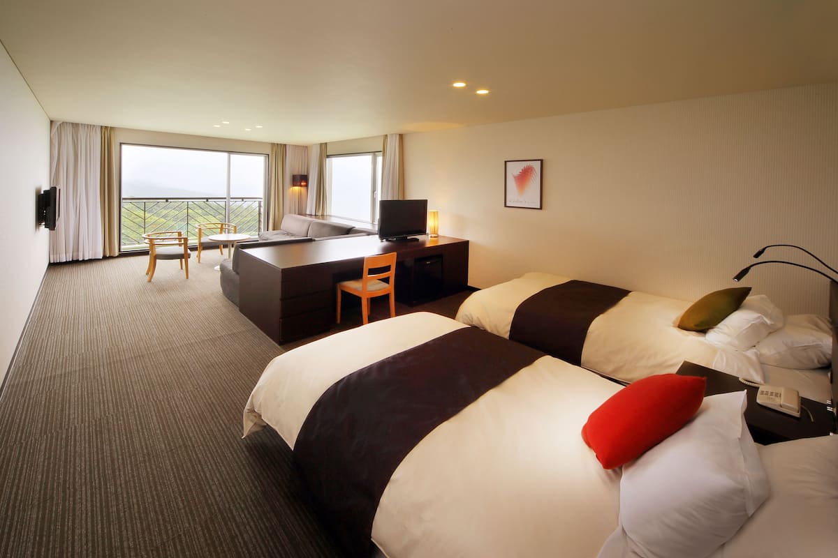 A hotel room with two beds, a desk, a chair, a TV, and a large window with curtains.