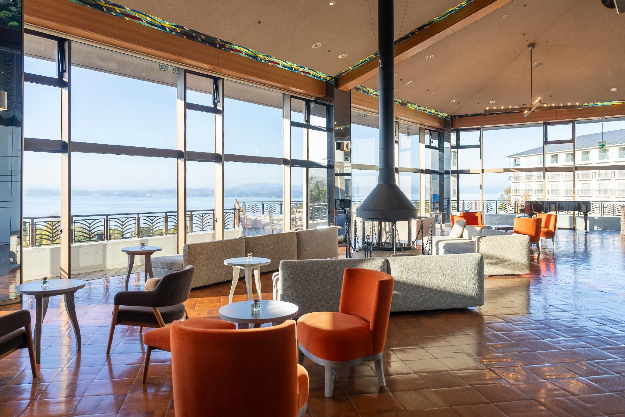 Amplio espacio interior con grandes ventanales que ofrecen vistas al mar, amueblado con asientos naranja y gris, y un techo alto con una lámpara central.