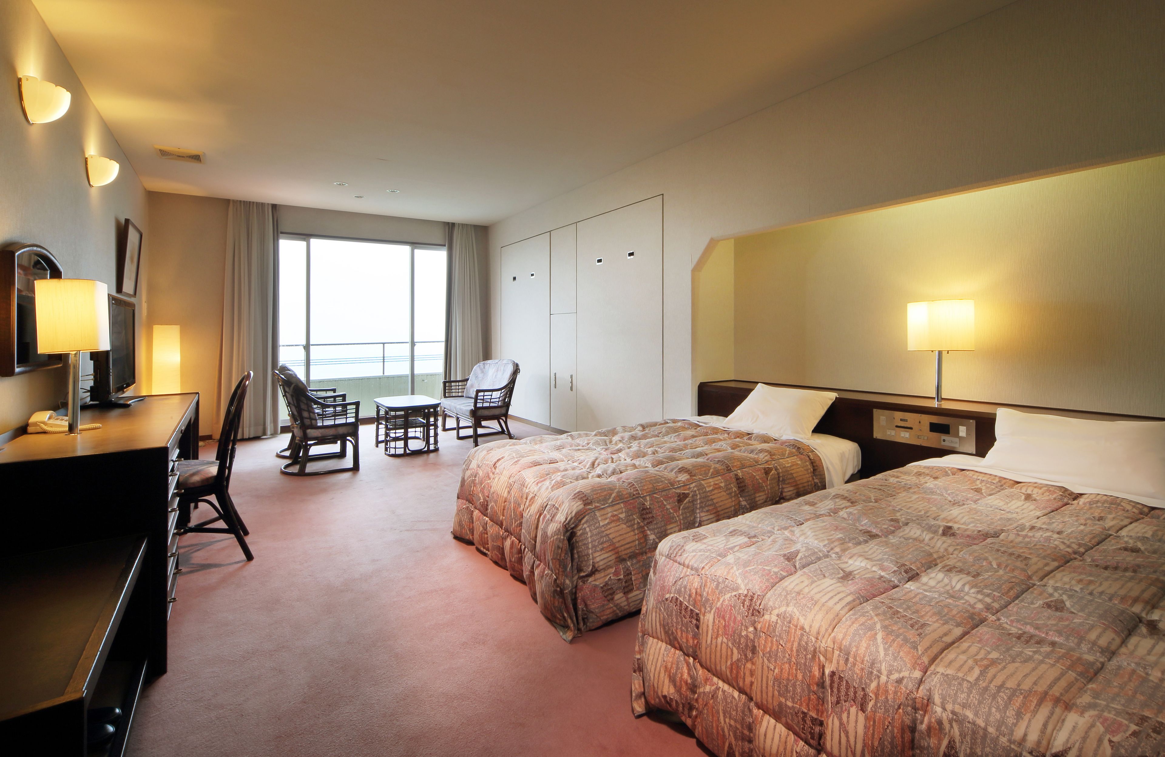 A hotel room with two beds, a desk, a chair, a TV, and a large window.