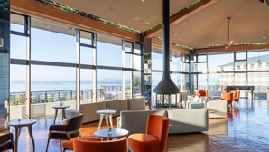Een ruime binnenzone met grote ramen met uitzicht op de zee, ingericht met oranje en grijze zitplaatsen, en een hoog plafond met een centraal hanglamp.