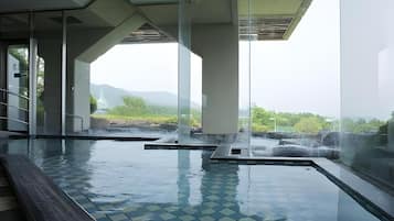 An indoor swimming pool with clear glass walls, a tiled floor, and a view of greenery and mountains in the background.