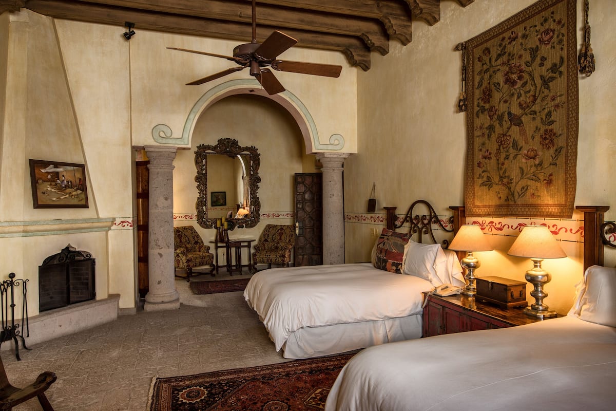 A hotel room with two beds, a ceiling fan, a large floral tapestry, and a stone fireplace.