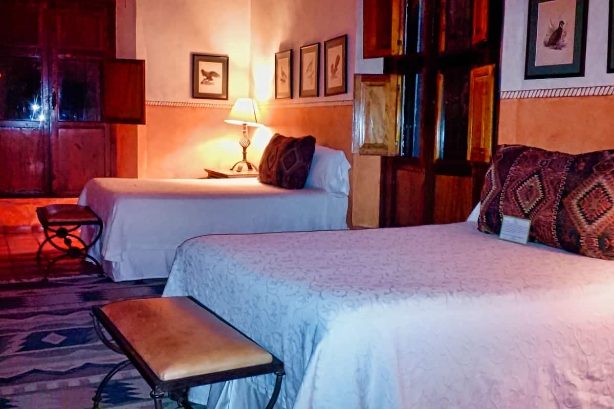 A hotel room with two beds, a bench, a table, and a lamp.
