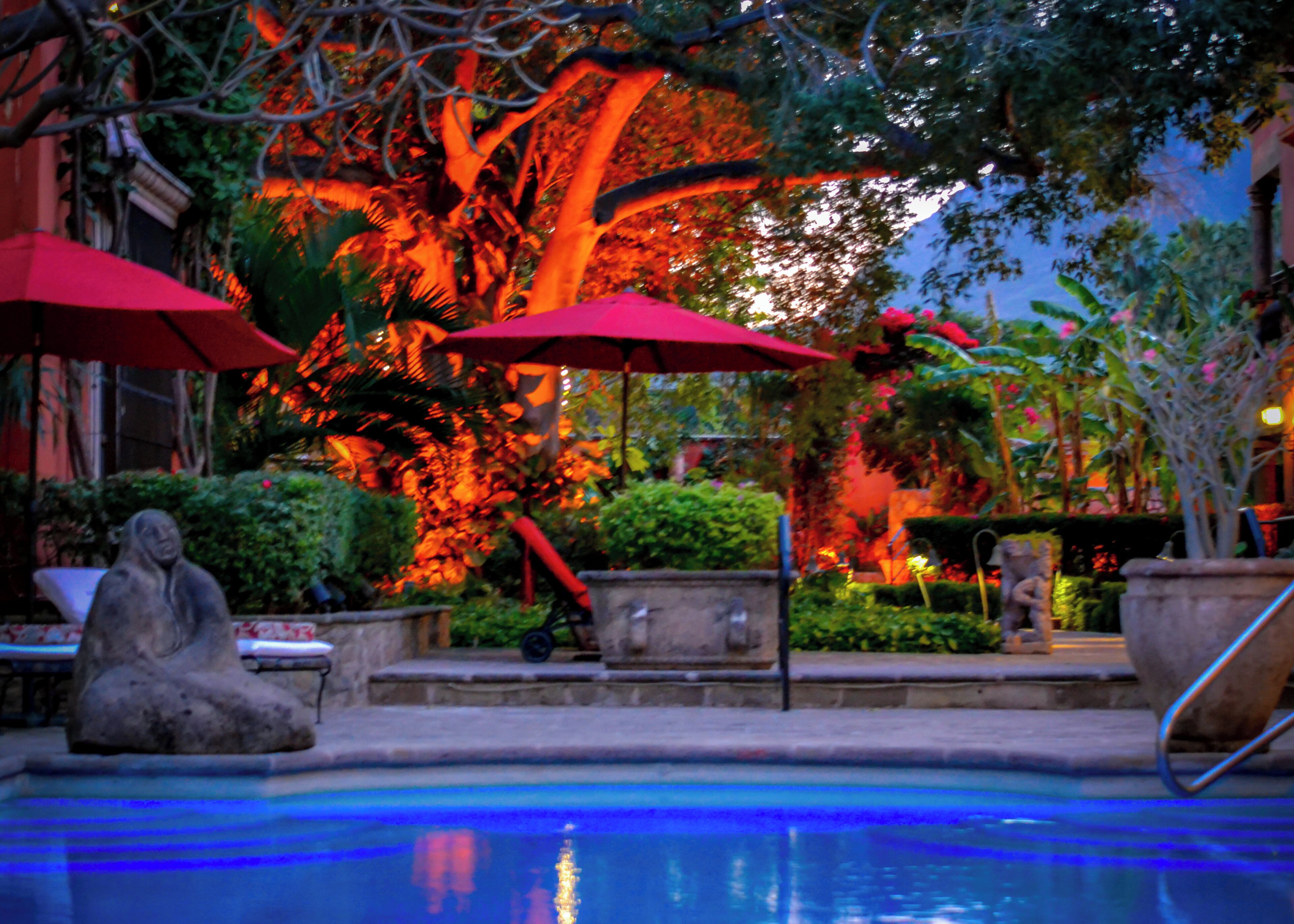 Zona de piscina con sombrillas rojas, estatuas de piedra y abundante vegetación.