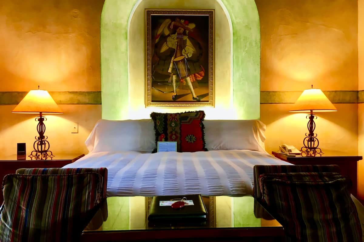 A hotel room with a bed, two lamps, a painting above the bed, and two chairs.