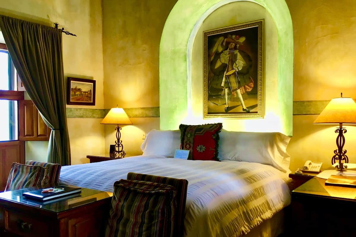 A hotel room with a bed, two bedside tables, a desk with a chair, a lamp, a painting above the bed, and a window with curtains.