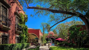 A pathway leading to a building with a red facade and arched entrance, surrounded by lush greenery and a clear blue sky. - Hacienda de los Santos (Alamos)