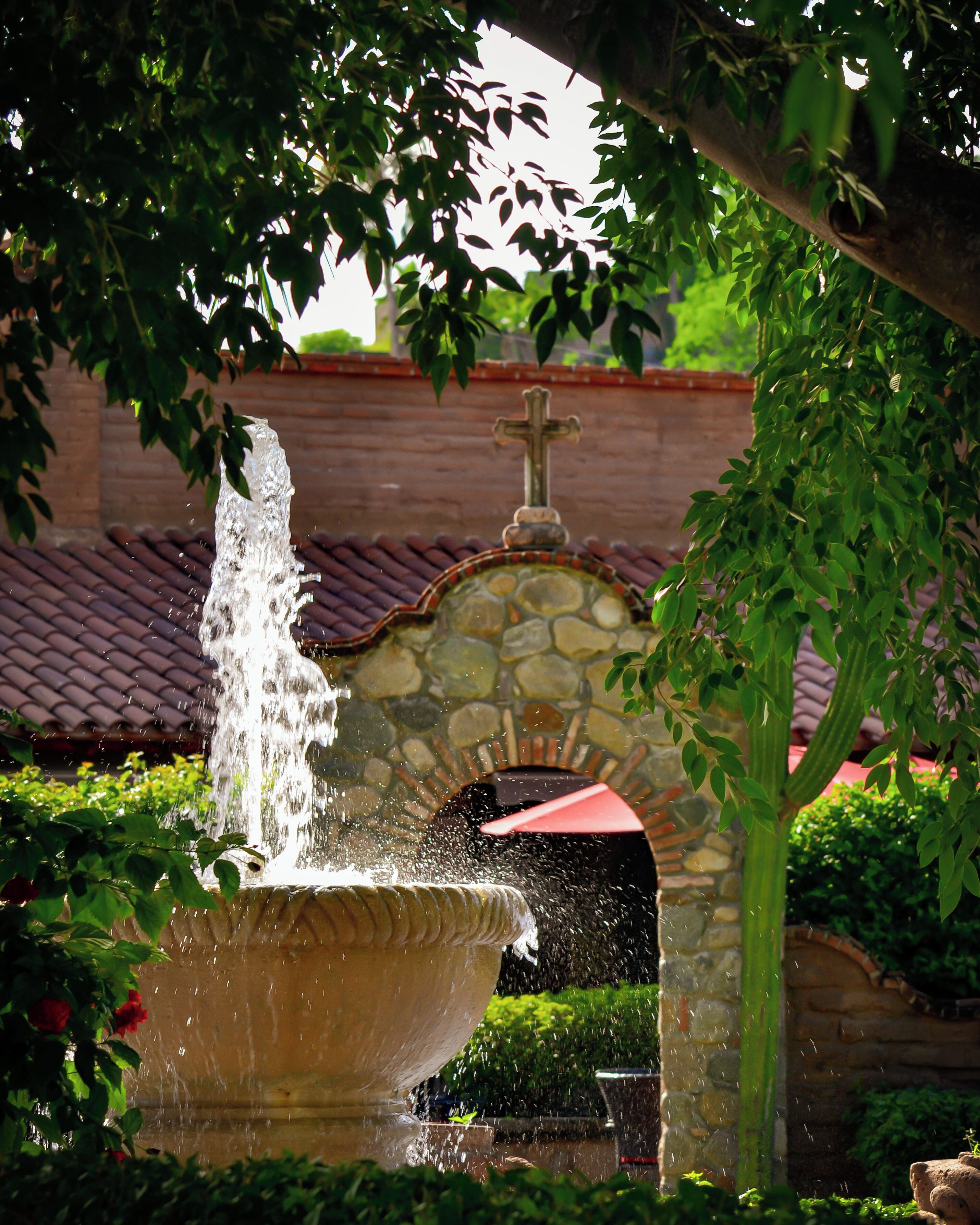 A stone fountain with water spouting from a central column, surrounded by lush greenery and a stone archway.