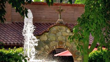 A stone fountain with water spouting from a central column, surrounded by lush greenery and a stone archway.