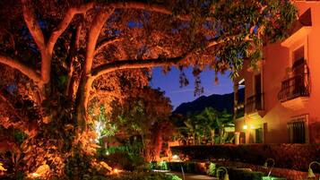 A nighttime view of a building with a large tree in the foreground, a well-lit pathway, and a mountain in the background.