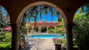An outdoor pool area with a clear view of a building with red walls and arches, surrounded by greenery and potted plants. - Hacienda de los Santos (Alamos)