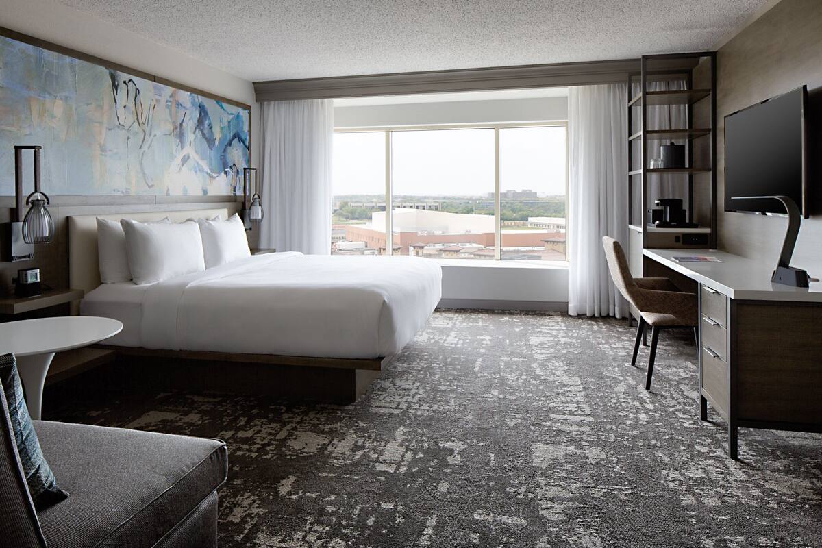 A hotel room with a large bed, a desk, a chair, a TV, and a view of the city through large windows.