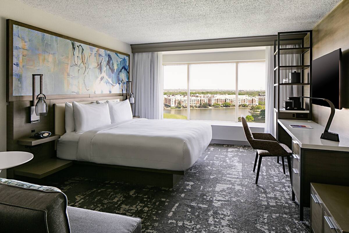A hotel room with a large bed, a desk, a chair, a TV, a bookshelf, and a painting on the wall.